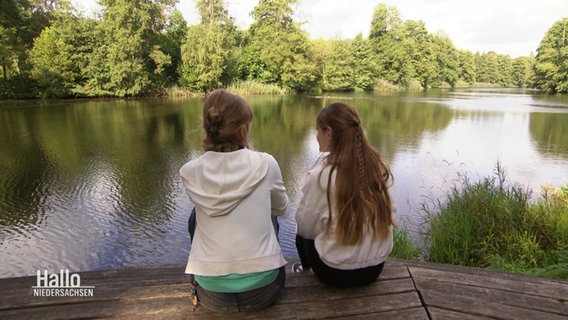 Zwei junge Frauen sitzen nebeneinander und schauen auf einen See. © Screenshot 