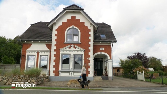 Eine Frau mit zwei Hunden geht auf einem Bürgersteig spazieren, im Hintergrund steht ein altes Backsteinhaus. © Screenshot 