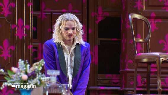 Ein Schauspieler steht in schillerndem lila Anzug auf der Bühne. © Screenshot 