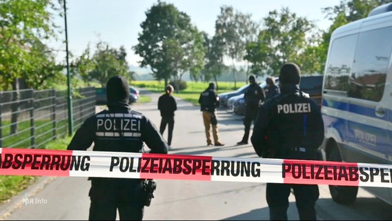 Polizisten und Einsatzfahrzeuge hinter einer Polizeiabsperrung. © Screenshot 