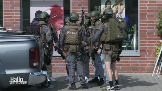 Maskierte und durch ihre Ausrüstung gut geschützte Spezialeinsatzkräfte der Polizei stehen in einer kleiner Gruppen auf dem Bürgersteig. © Screenshot 