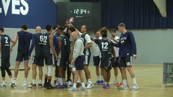 Eine Basketballmannschaft klatscht sich auf dem Feld in einer Sporthalle in der Mitte eines Kreises ab. © Screenshot 