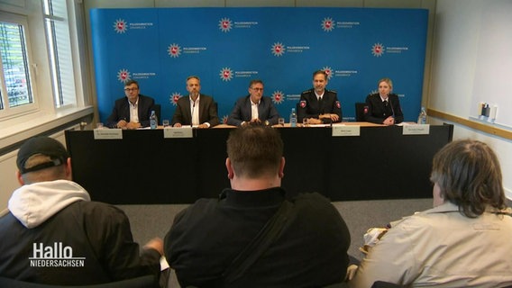 Bei einer Pressekonferenz der Polizei Niedersachsen sitzen mehrere Männer in Anzug und Uniform an einem langen Tisch vor einer Logo-Wand. © Screenshot 