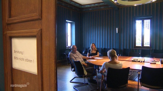 Zwei Frauen und ein Mann sitzen an einem größeren Tisch in einem Sitzungssaal beratend zusammen. © Screenshot 