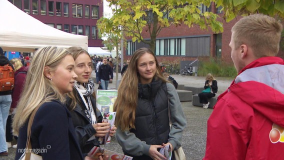 Drei jüngere Frauen hören einem Mann in roter Jacke auf einem Universitätscampus zu. © Screenshot 