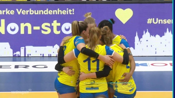 Die Volleyballerinnen des SSC Palmberg Schwerin stehen bei einem Spiel in einem engen Kreis mit um die Schultern gelegten Armen. © Screenshot 