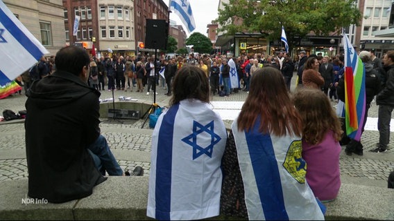 Auf einem öffentlichen Platz bekunden mehrere Menschen bei einer Demonstration ihre Solidarität mit Israel. Im Vordergrund sitzen drei kleiner Kind mit um den Oberkörper geschlungenen israelischen Flaggen. © Screenshot 