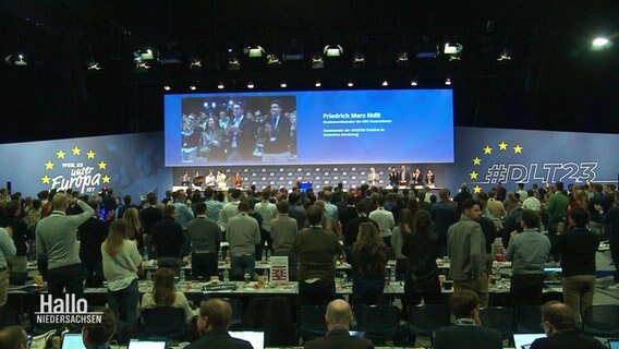Bei einer Generalversammlung der Jungen Union stehen viele Zuhörende bei einer Rede von Friedrich Mertz, der von Ihnen auf einer Bühne steht. © Screenshot 