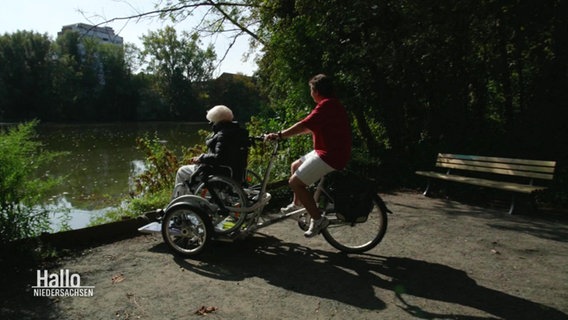 Eine Frau fährt eine ältere Dame auf einem Lastenrad-ähnlichen Fahrrad an das Ufer eines Sees. © Screenshot 