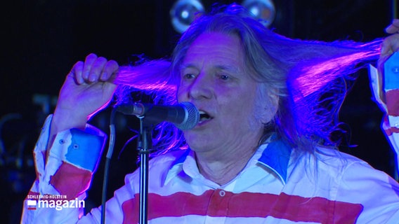 Ein Sänger zieht sich die langen Haare wie Zöpfe zu beiden Seiten während er in ein Standmikrofon singt. © Screenshot 