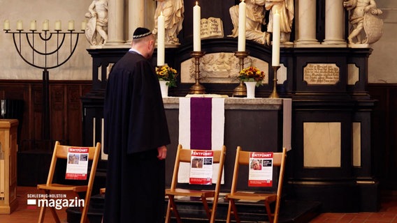 Ein jüdischer Rabbiner steht vor einem christlichen Altar vor Stühlen auf denen Plakate stehen. © Screenshot 