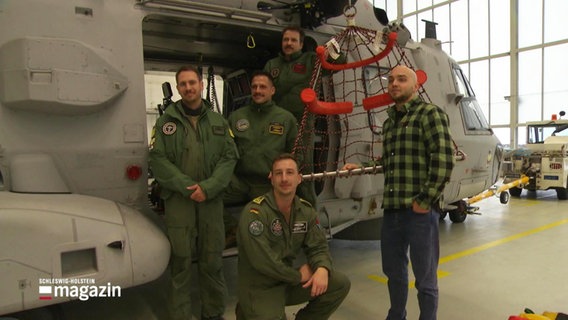 Ein junger Mann in Alltagsklamotten und mehrere Männer in Uniform stehen vor einem Helikopter. © Screenshot 