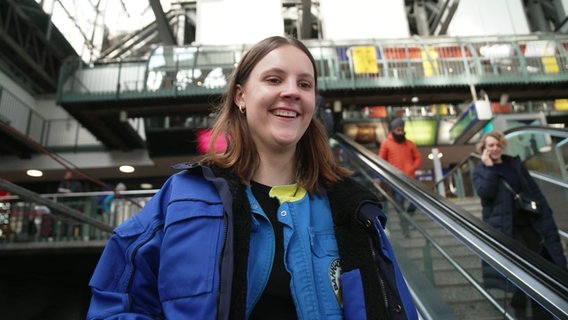 Eine junge Frau in einem Bahnhof lacht in die Kamera. © Screenshot 