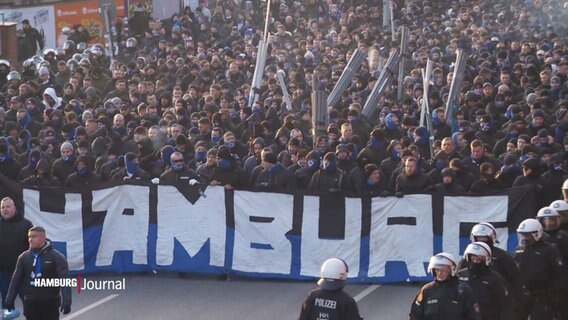 Fanblock des HSV beim Anmarsch zum Millertor-Stadion. Vorne wird ein Banner mit dem Schriftzug "Hamburgin den Farben des Vereins getragen, Polizisten flankieren den Fanblock. © Screenshot 