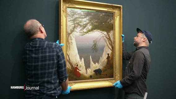 Zwei Männer mit blauen Gummihandschuhen hängen vorsichtig ein goldgrahmtes Gemälde von Maler Caspar David Friedrich an die Wand. © Screenshot 
