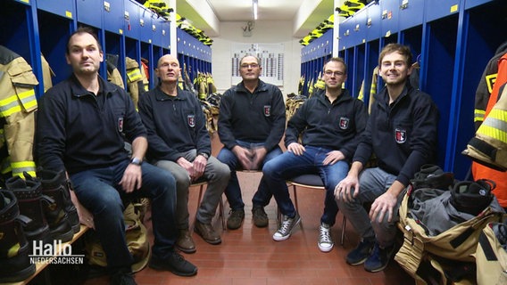Mitglieder der Familie Köster mit Ortsbrandmeister Thomas Köster aus Achim sitzen im Halbkreis vor ihren Spinden mit den Feuerwehruniformen. © Screenshot 