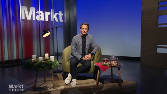 Moderator Jo Hiller sitzt im Studio von "Markt" auf einem Sessel, rechts neben ihm ein Tischchen mit Tee und Keksen, links von ihm ein Tischchen mit einem Adventskranz mit weißen Kerzen; die erste Kerze brennt. © Screenshot 
