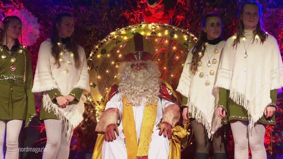 Der Nikolaus sitzt auf einer Bühne. Seine Helferinnen stehen links und rechts von ihm. © Screenshot 