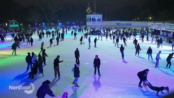 Die Eisbahn in Hamburg Planten und Blomen bei Nacht. Viele Menschen tummeln sich auf dem Eis und bunte Lichter erstrecken sich über das Eis. © Screenshot 