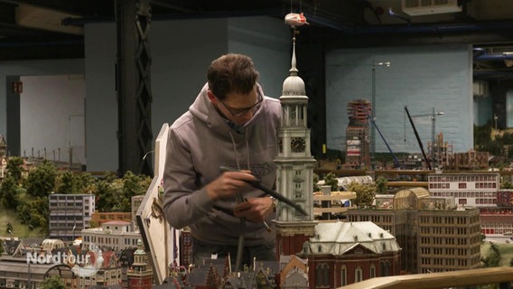 Ein Mann saugt das Modell des Hamburger Michels im Miniatur Wunderland ab. © Screenshot 