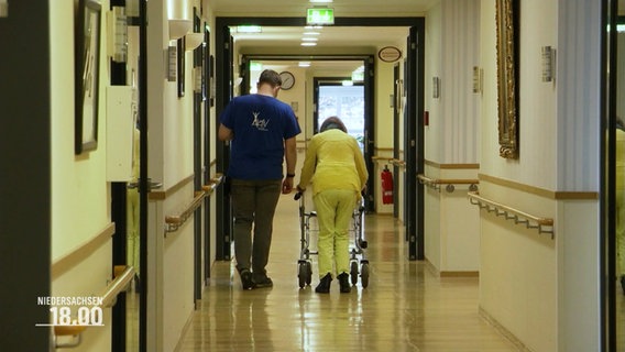 Ein Pfleger und eine Frau mit Rollator auf einem Flur eines Pflegeheimes. © Screenshot 