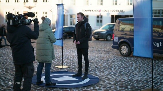 Auf einem Platz liegt ein runder Teppich mit dem ARD-Logo, darauf steht ein Mann in einer Interview-Situation: Eine Reporterin stellt Fragen, hinter ihr steht ein Kameramann, jemand hält ein Mikrofon. © Screenshot 