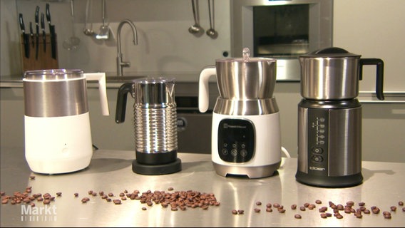 Verschiedene Milchaufschäumer stehen nebeneinander auf einer Edelstahl-Arbeitsfläche, im Vordergrund Kaffeebohnen. © Screenshot 