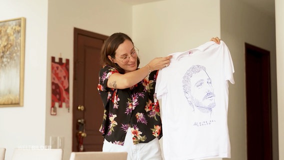 Eine Frau hält ein T-shirt mit dem Portrait ihres verstorbenen Mannes in die Kamera. © Screenshot 