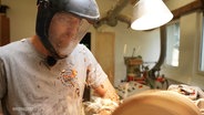 Ein Mann verarbeitet Holz und trägt dabei einen Gesichtsschutz. © Screenshot 