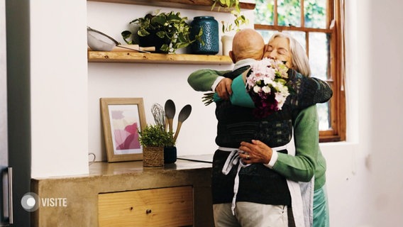Eine ältere Dame und ein älterer Herr umarmen sich. Sie hält dabei einen Blumenstrauß in der Hand. © Screenshot 
