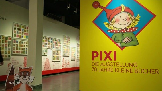 Im Vordergrund ein Plakat mit dem Logo der Pixi-Bücher und darunter die Aufschrift: "PIXI - Die Ausstellung. 70 Jahre kleine Bücher". Im Hintergrund sieht man Teile der Ausstellung. © Screenshot 