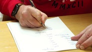 Eine Person schreibt Einkommenskalkulationen auf einen Zettel. © Screenshot 