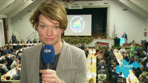 Die Reporterin Barbara Kreuzer berichtet aus einer Halle in Jork. Viele Menschen sitzen hinter ihr an langen Tischen vor einer Bühne. © Screenshot 