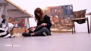 Simin Jalilian sitzt auf dem Boden eines Ateliers, im Hintergrund sind zwei ihrer Werke zu sehen. © Screenshot 
