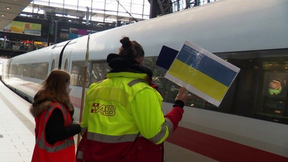 Eine Frau steht mit einem Ukraine-Schild am Bahnhof an einem Zug. © Screenshot 