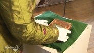 Eine Frau greift mit Stoffhandschuhen nach einem sehr alten Buch, das auf einem Sockel liegt. © Screenshot 