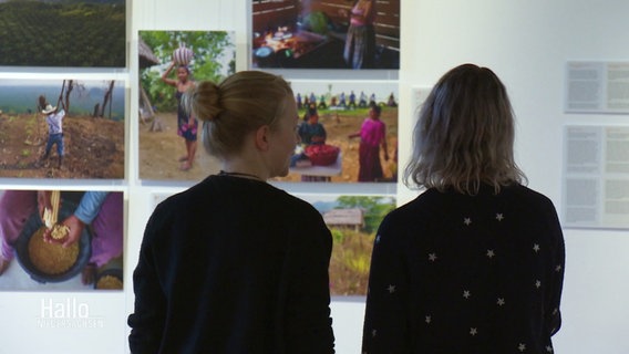 Zwei Frauen schauen sich eine Wand mit Fotos an. © Screenshot 
