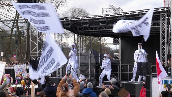 Die Hamburger Band Deichkind spielt auf einer Demo gegen Rechtsextremismus am 25.02.2024. Sie tragen weiße Anzüge und schwenken weiße Fahnen, auf beidem steht etwas geschrieben. © Screenshot 