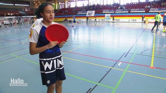 Ein Mädchen steht mit einer roten Frisbee in der Hand in einer Sporthalle © Screenshot 
