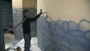 Ein Mann malt mittels Airbrush eine graue Steinmauer an eine Innenwand. © Screenshot 