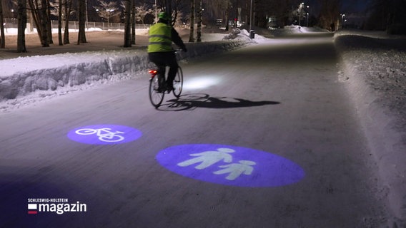 Auf einem verschneiten, aber geräumten Fahrrad- und Gehweg leuchten blau die Smybole für den Fahrrad- und Fußgängerstreifen. Ein winterfest gekleideter Mann fährt auf einem Fahrrad. © Screenshot 