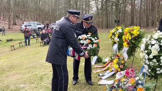 Zwei Polizeibeamte legen einen Gedenkkranz zu weiteren Blumengestecken. © Screenshot 
