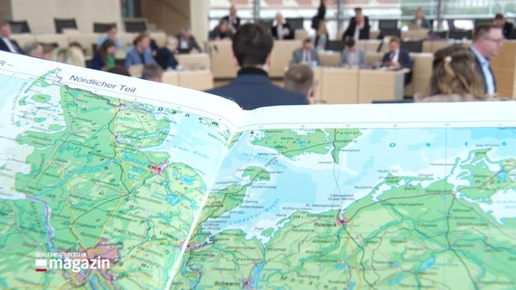 Hinter einem Atlas mit Ostseekarte sieht man Mitglieder des Landtags während ihrer Tagung. © Screenshot 