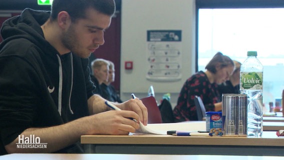 Ein Schüler während der Abiturprüfung an einem Einzeltisch, vor ihm liegen auch Snacks und Getränke. © Screenshot 