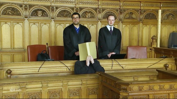 Zwei Menschen in Roben stehen über einer Person, die sich eine Mappe vor ihr Gesicht hält in einem Gerichtssaal. © Screenshot 