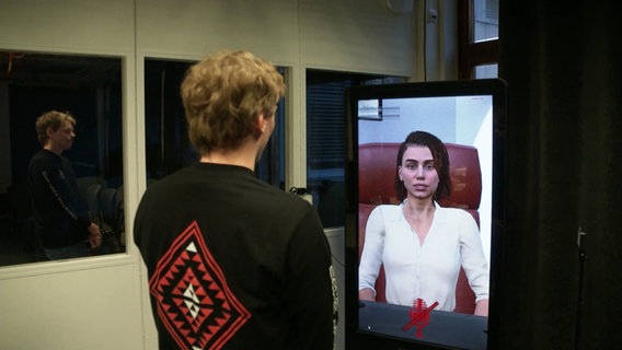 Ein Mann steht vor einem Bildschirm, auf dem eine compter-animierte Frau zu sehen ist. © Screenshot 