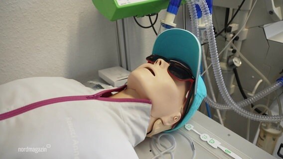Eine Puppe als Dummy, angezogen mit einer Sweatjacke, Cappie und Sonnenbrille, liegt auf einer Lige. Im Hintergrund medizinische Geräte. © Screenshot 