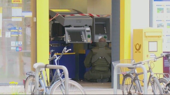 Eine Person in dunkelgrüner Schutzkleidung hockt vor einem abgesperrten Geldautomaten. © Screenshot 