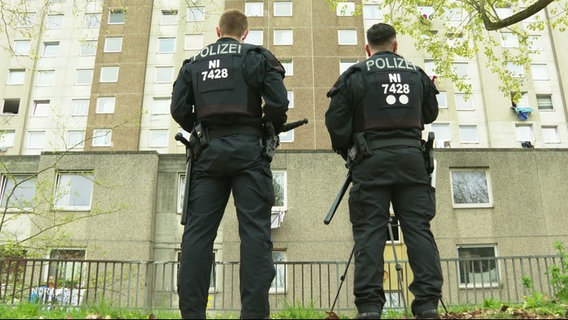 Aus der Froschperspektive: Zwei mit Schutzkleidung, Schlagstöcken und Waffen ausgerüstete Polizeibeamte stehen vor einem Plattenbau. © Screenshot 
