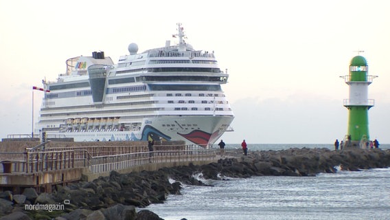 Die "AIDAmar"  fährt auf Warnemünde zu. Daneben ragt ein grün-weißer Leuchtturm vom Pier empor. © Screenshot 
